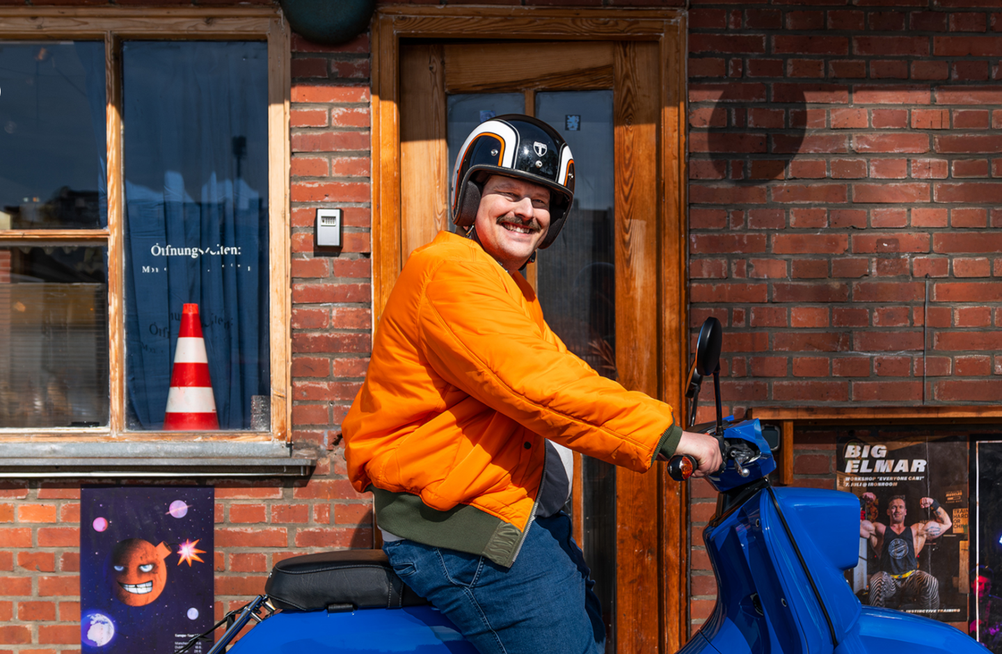 Ein Mann mit oranger Jacke sitzt auf einem blitzblauen Roller und lächelt zufrieden.